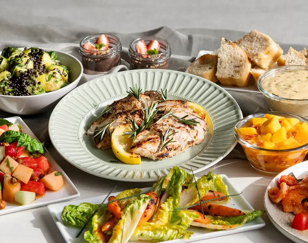 Sommarbuffé för catering med grillad kyckling, melon- och salladsrätter, bröd, såser, dessert i glas och färgstarka tillbehör upplagda på ett buffébord.