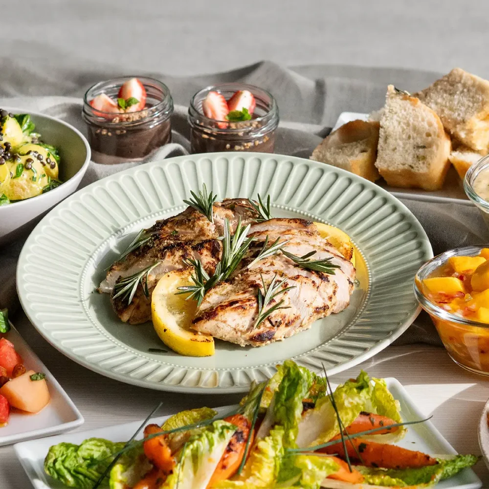 Sommarbuffé för catering med grillad kyckling, melon- och salladsrätter, bröd, såser, dessert i glas och färgstarka tillbehör upplagda på ett buffébord.