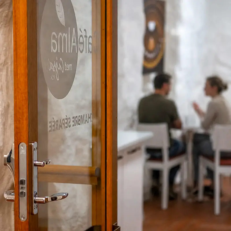 Två cafégäster samtalar vid ett bord i Café Almas chambre séparée, sedda genom den öppna glasdörren.